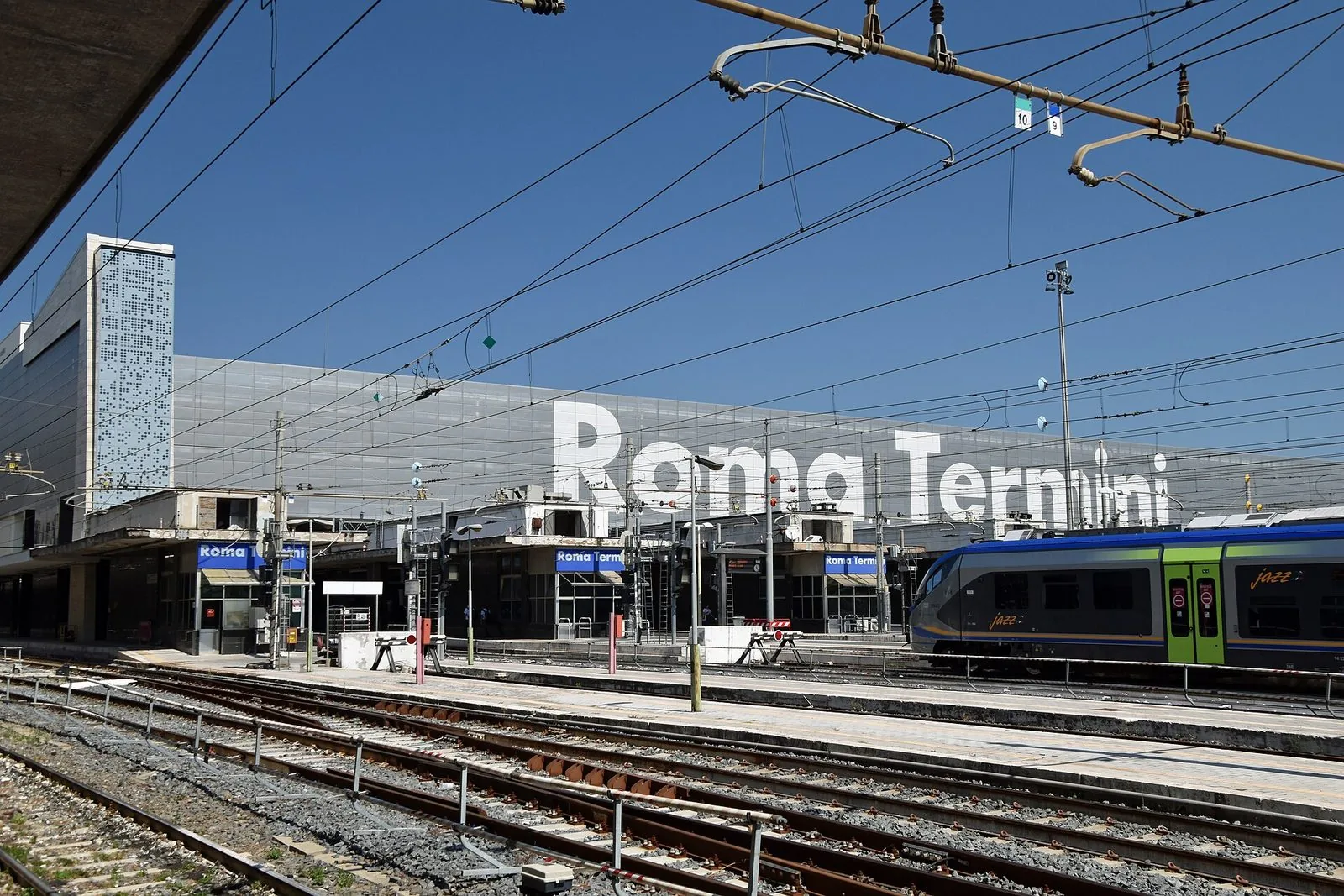 La stazione Roma Termini vista dai binari: la lunga facciata con la grande scritta Roma Termini, la torre chiara a sinistra, un treno regionale in sosta e le linee aeree sotto il cielo azzurro