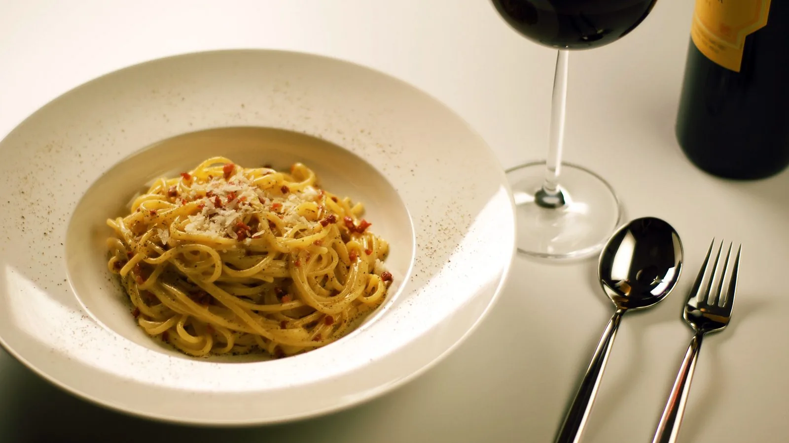 Un piatto fondo bianco con spaghetti alla carbonara, pezzetti di guanciale, pecorino grattugiato e pepe nero; accanto un calice di vino rosso, una bottiglia, un cucchiaio e una forchetta su una tovaglia chiara