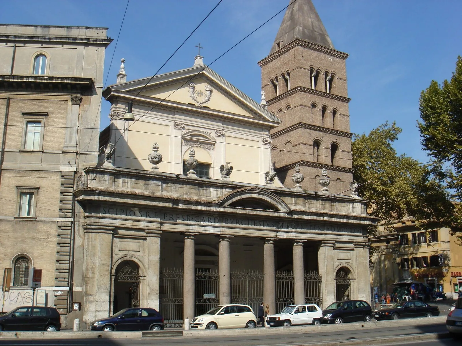 Facciata della basilica di San Crisogono a Trastevere con il portico a colonne, il timpano bianco e il campanile romanico in mattoni, auto parcheggiate davanti e cielo azzurro