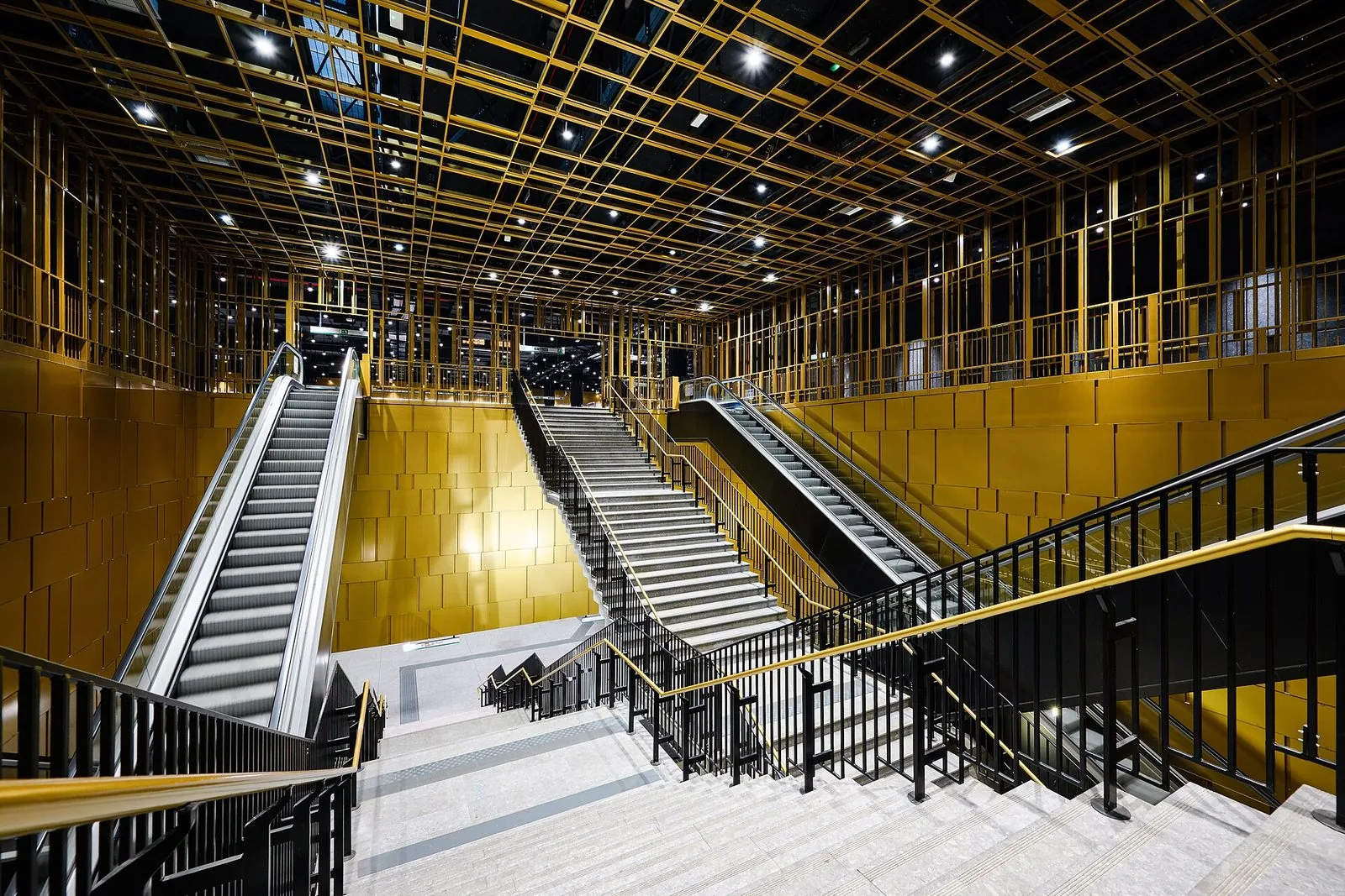 L'atrio della stazione Colosseo della Metro C: scale fisse e scale mobili che scendono verso i binari, pareti rivestite di pannelli dorati e un soffitto a griglia metallica color oro con faretti