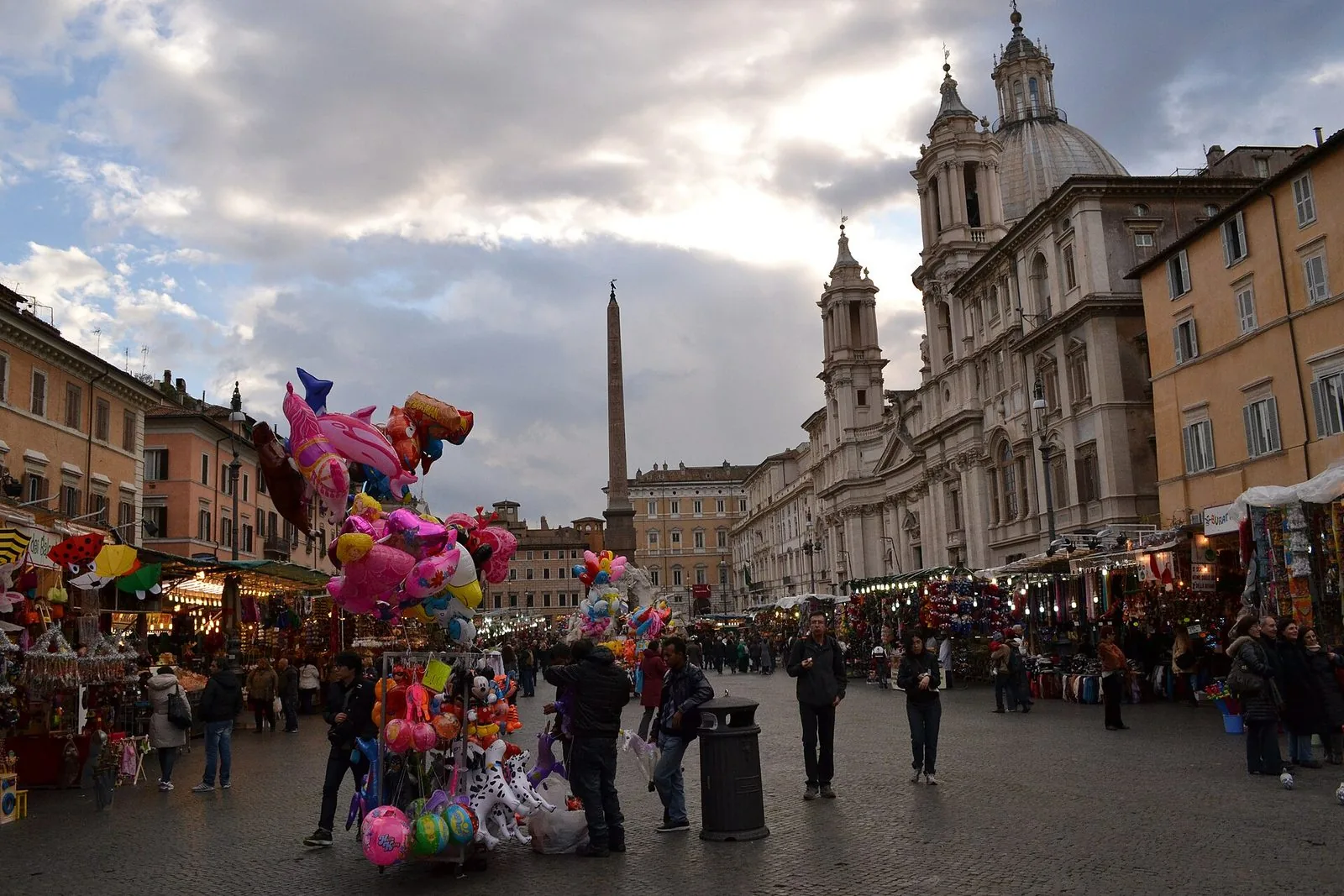 Piazza Navona con file di bancarelle e venditori di palloncini colorati, l'obelisco della Fontana dei Quattro Fiumi e la facciata con le torri campanarie di Sant'Agnese in Agone sotto un cielo nuvoloso