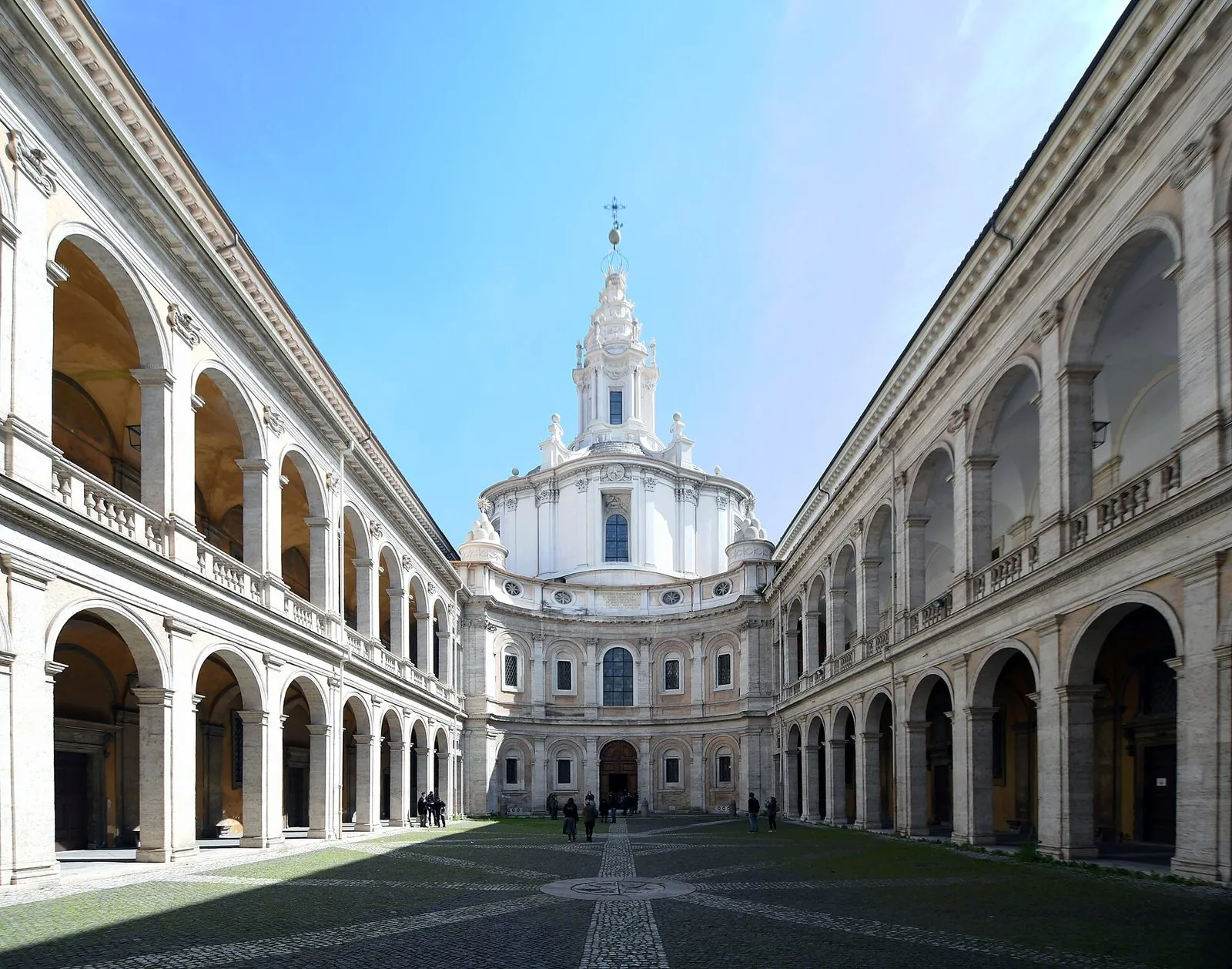 Il cortile porticato del Palazzo della Sapienza a Roma con due ordini di arcate ai lati e, sul fondo, la facciata concava di Sant'Ivo alla Sapienza sormontata dalla cupola bianca e dalla lanterna a spirale, sotto un cielo azzurro