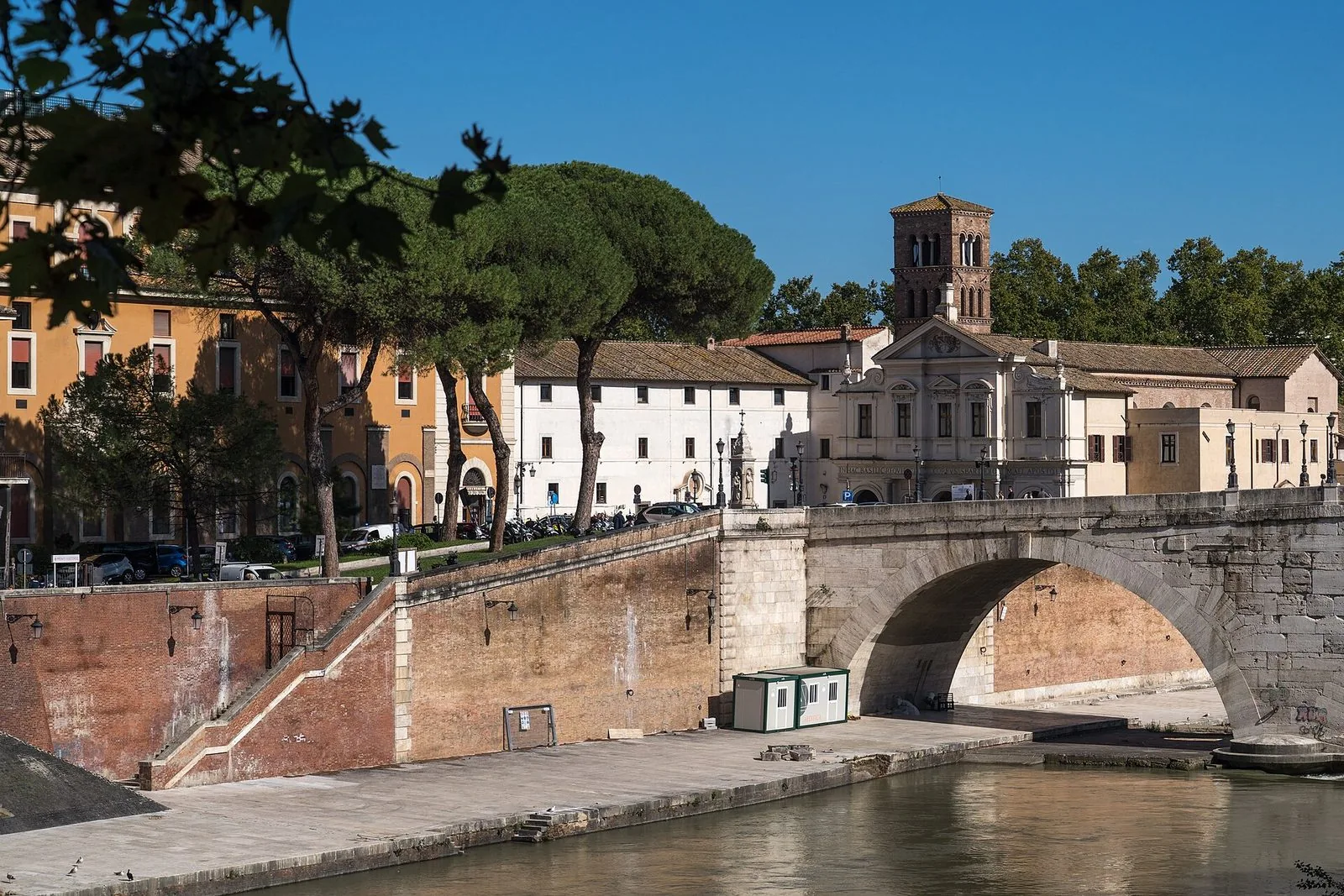 L'Isola Tiberina vista dalla riva del Tevere in una giornata di sole: un ponte romano ad arco in pietra, i pini sopra il muraglione in mattoni, la facciata chiara della basilica di San Bartolomeo con il campanile romanico e i palazzi color ocra