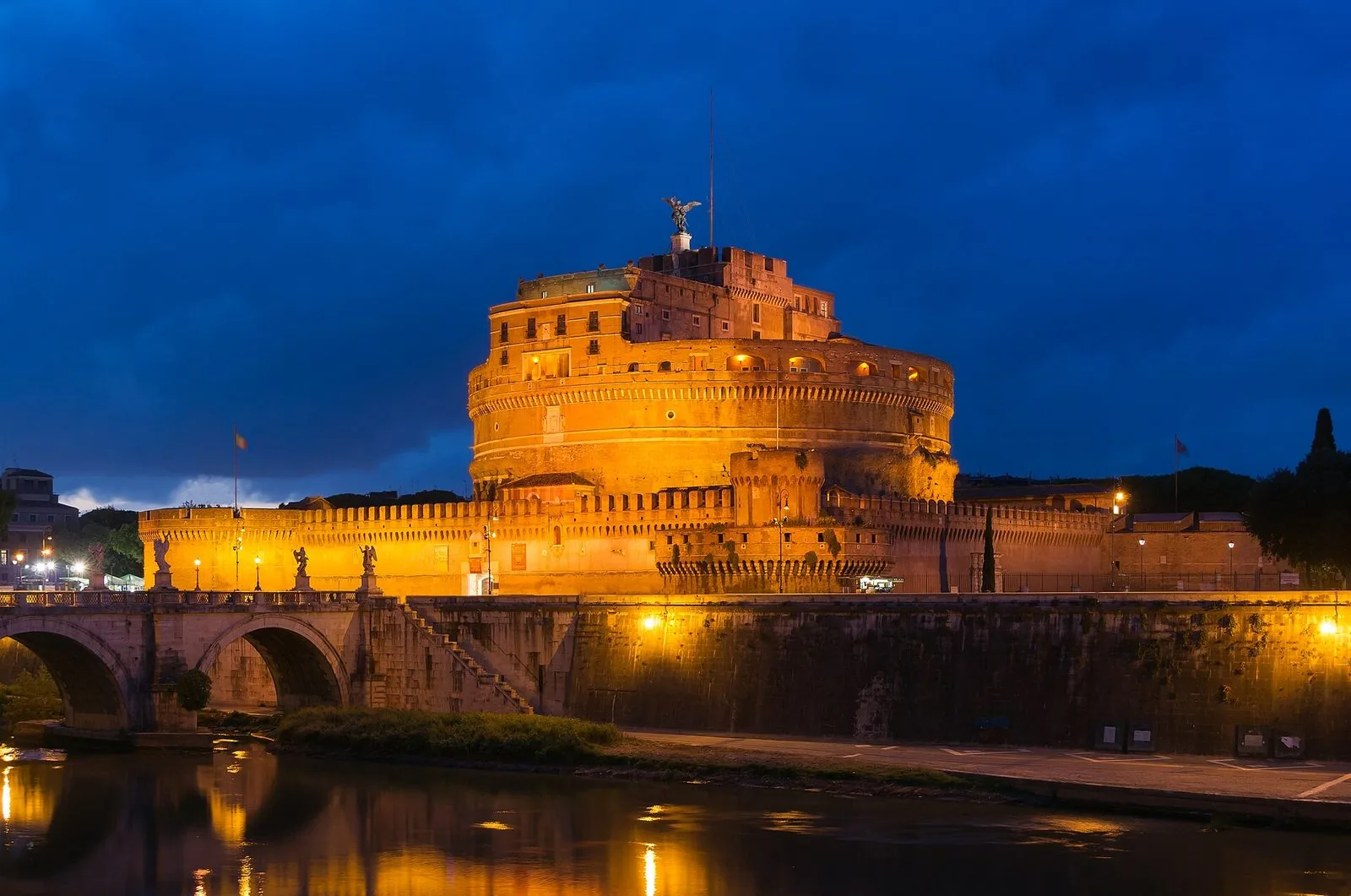 Castel Sant'Angelo illuminato al crepuscolo sotto un cielo blu intenso, con l'angelo in bronzo in cima, le mura merlate, il Ponte Sant'Angelo con le statue a sinistra e il riflesso delle luci nel Tevere