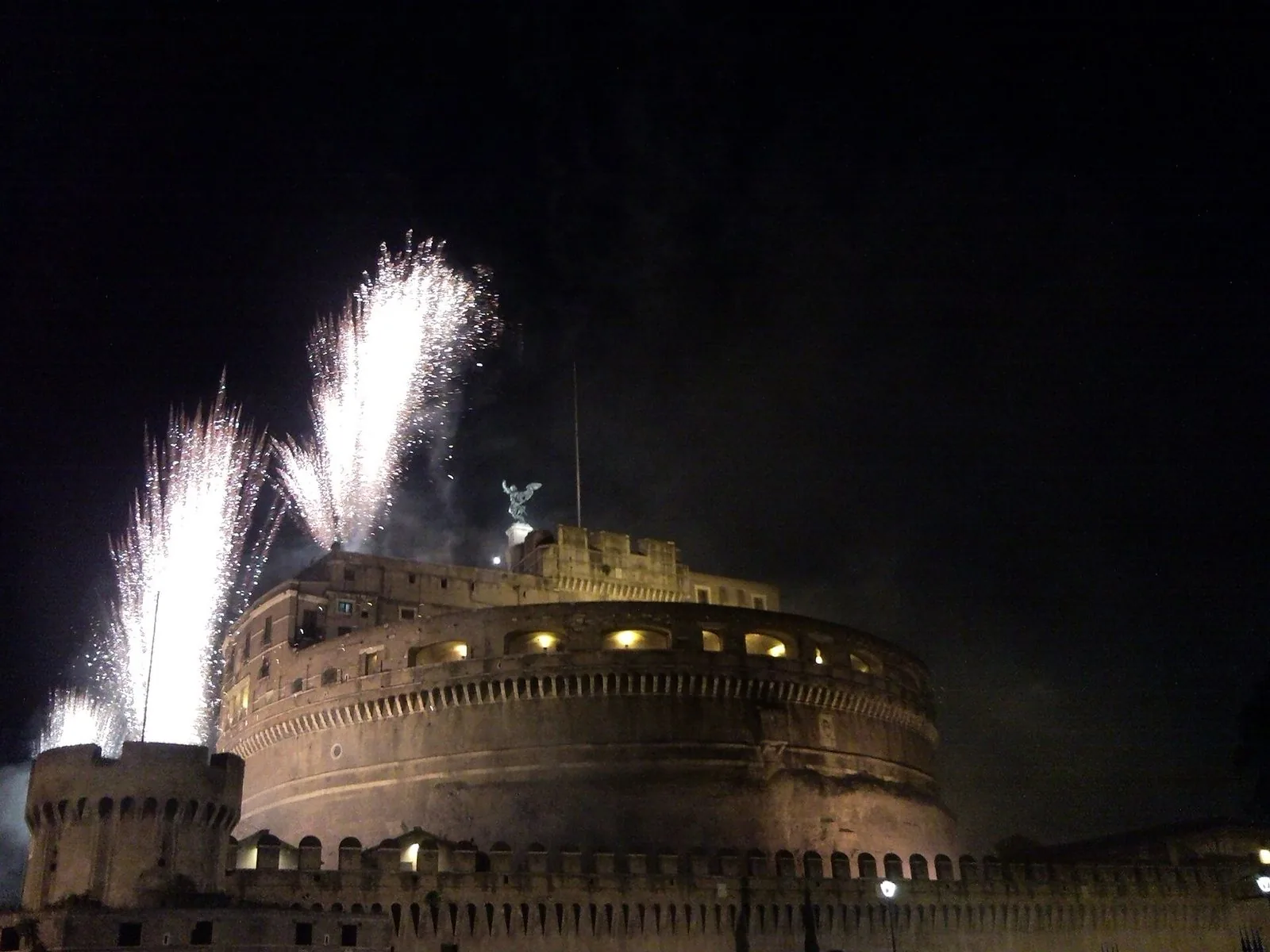 Castel Sant'Angelo illuminato di notte durante la Girandola, con due getti di fuochi d'artificio bianchi che si alzano sopra la mole cilindrica e il torrione merlato in primo piano