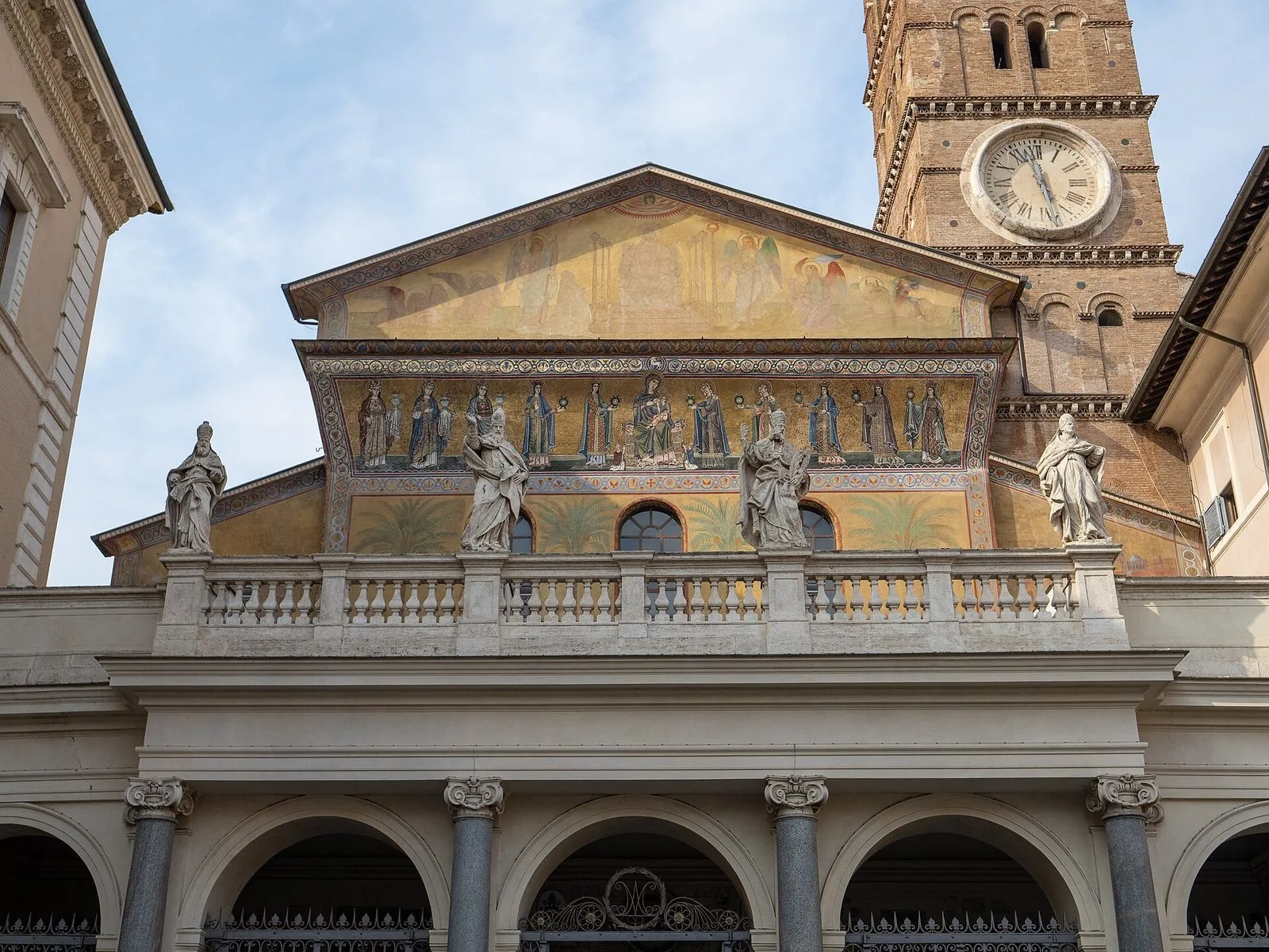 Facciata della basilica di Santa Maria in Trastevere con il fregio a mosaico dorato, le statue di santi sulla balaustra sopra il portico e il campanile romanico con l'orologio, sotto un cielo azzurro
