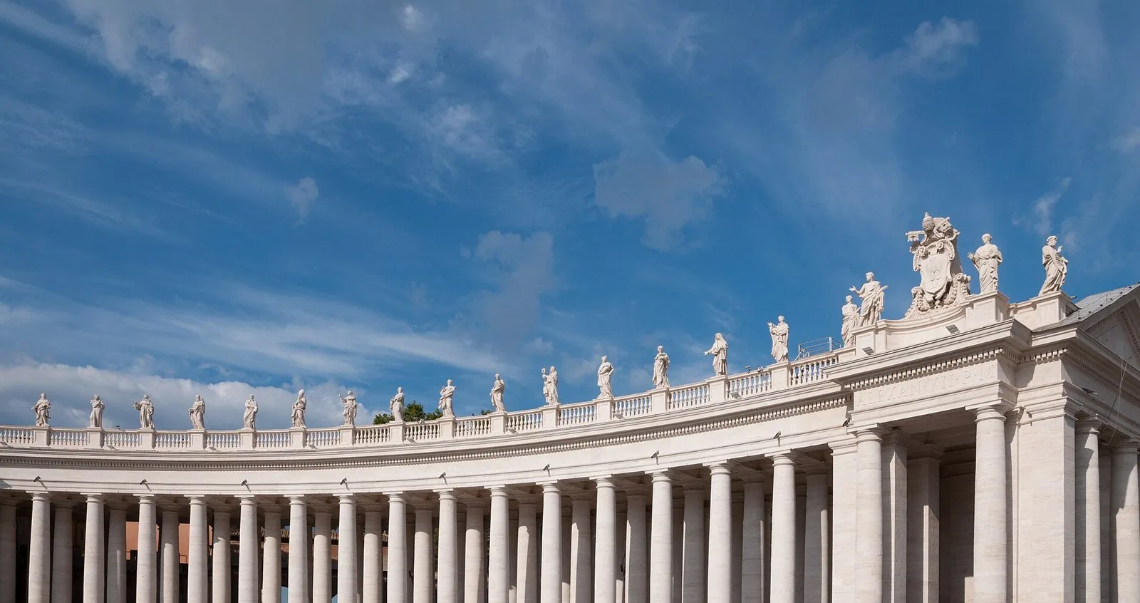 Il colonnato di Bernini in Piazza San Pietro sotto un cielo azzurro con nuvole leggere: file di colonne in travertino sormontate da una balaustra con statue di santi e da uno stemma papale scolpito